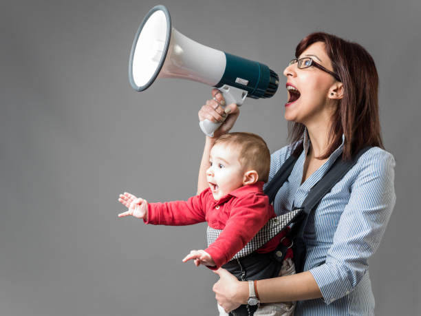 Young mother carrying her baby in baby carrier and shouting through megaphone in front of gray background. Baby is wearing a red t-shirt and he is shouting and excited too. Shot in studio with a medium format camera.